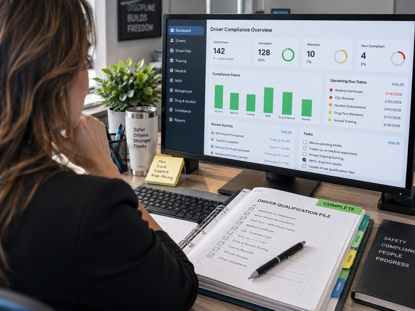 Over the shoulder of a safety director at her desk, the driver compliance dashboard on screen and an open driver qualification file in a binder in front of her, every line checked